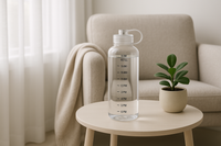 A sleek, modern home vignette styled like a high-end magazine editorial. Place a 1 L Tritan water bottle with time markings prominently on a minimalist side table. The scene includes a neutral-toned cozy armchair with a soft throw, a small plant in a ceramic pot, and gentle natural light filtering through sheer curtains. The atmosphere is fresh, healthy, and aspirational—showcasing the water bottle as a daily hydration essential that fits effortlessly into a chic, wellness-focused lifestyle